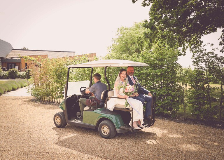 A bride and her dad travelling on the back of a golf cart to the wedding ceremony on a wedding day at Little Green Wedding Barn photographed by suffolk wedding photographer Hayley Denston Photography