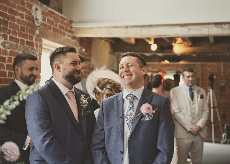 A groom and his bestman laughing together at the top of the aisle before a wedding ceremony on a wedding day at Little Green Wedding Barn photographed by suffolk wedding photographer Hayley Denston Photography