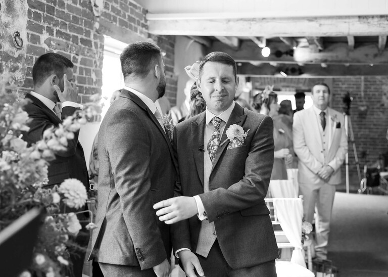 A black and white photo of a groom grabbing his bestman at the front of the ceremony as he feels emotional before the bride walks down the aisle on a wedding day at Little Green Wedding Barn photographed by suffolk wedding photographer Hayley Denston Photography