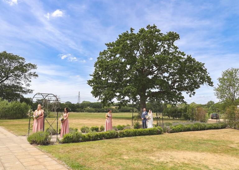 A bride and bridesmaids lined up to come in for a wedding ceremony outside with meadows behind them on a wedding day at Little Green Wedding Barn photographed by suffolk wedding photographer Hayley Denston Photography