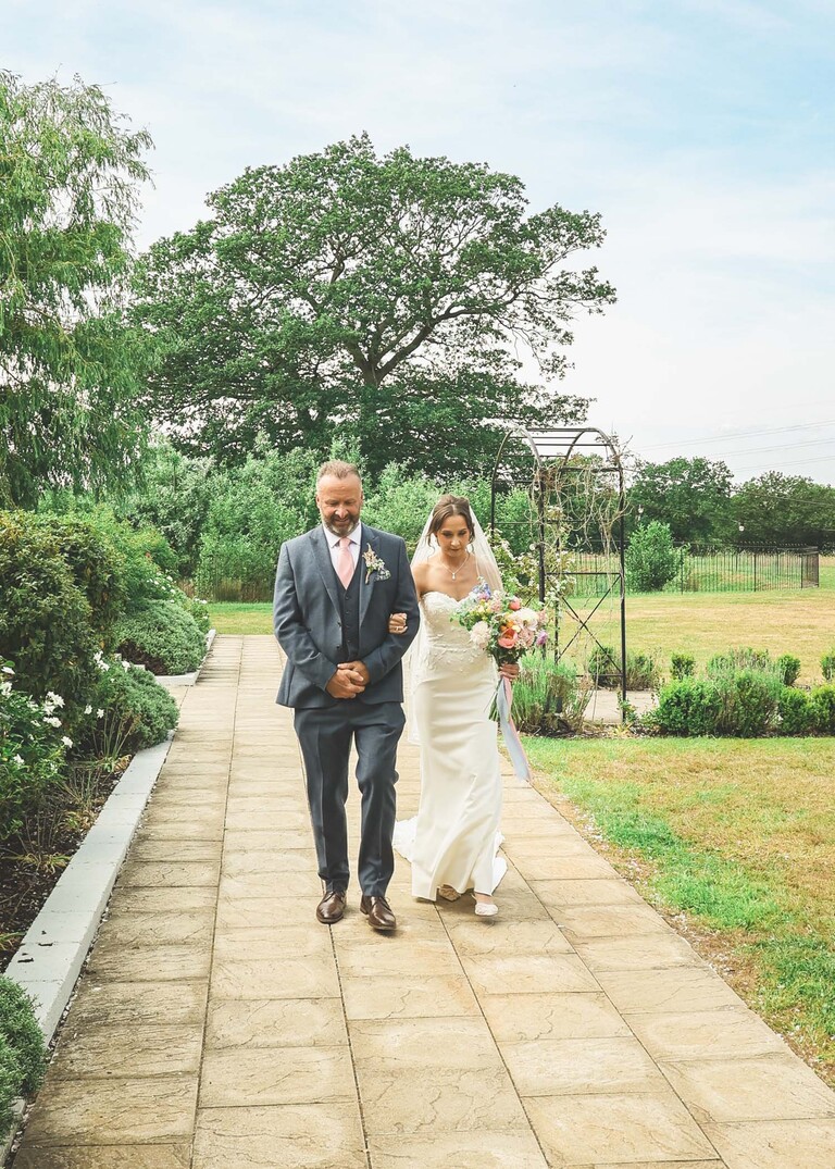 A bride and her dad walking down the path before entering for an indoor wedding ceremony on a wedding day at Little Green Wedding Barn photographed by suffolk wedding photographer Hayley Denston Photography