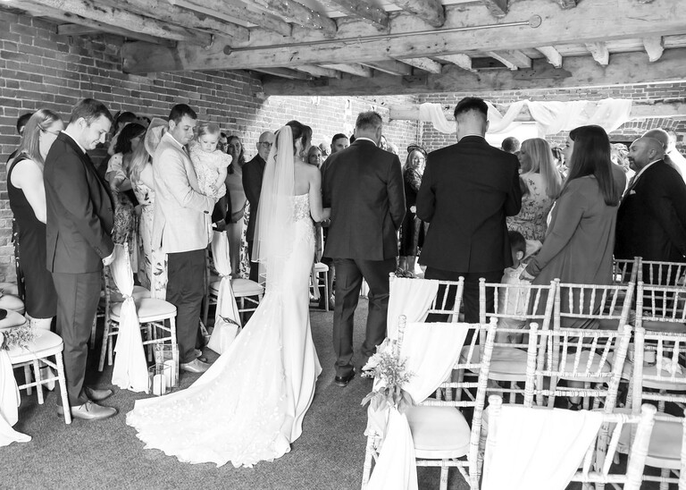 A black and white photo from behind of a bride and her dad walking up the aisle on a wedding day at Little Green Wedding Barn photographed by suffolk wedding photographer Hayley Denston Photography