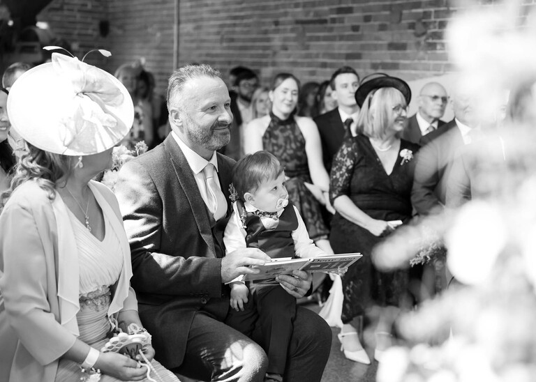 A black and white photo of a grandson sat on his grandads lap reading a book together whilst his mum and dad get married on a wedding day at Little Green Wedding Barn photographed by suffolk wedding photographer Hayley Denston Photography