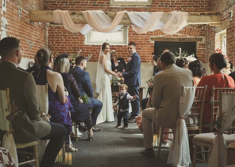 A bride and groom from the back of the ceremony room turning to face their son in the middle of the room during the wedding ceremony on a wedding day at Little Green Wedding Barn photographed by suffolk wedding photographer Hayley Denston Photography