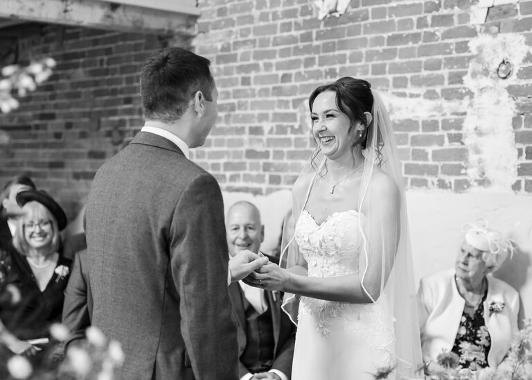 A bride putting the ring on the grooms finger during a wedding ceremony on a wedding day at Little Green Wedding Barn photographed by suffolk wedding photographer Hayley Denston Photography