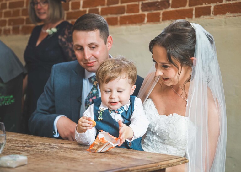 A pageboy sat on his parents laps eating cheddars as they sign the register on a wedding day at Little Green Wedding Barn photographed by suffolk wedding photographer Hayley Denston Photography