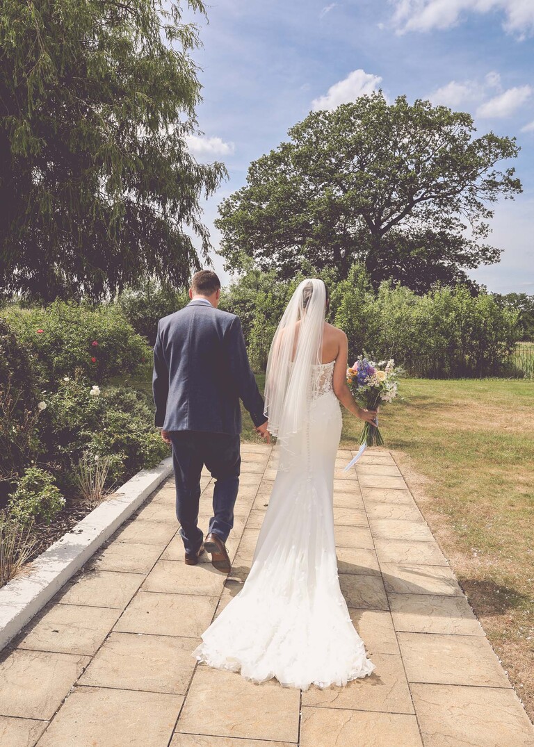 A bride and groom walking away down a path towards trees on a wedding day at Little Green Wedding Barn photographed by suffolk wedding photographer Hayley Denston Photography