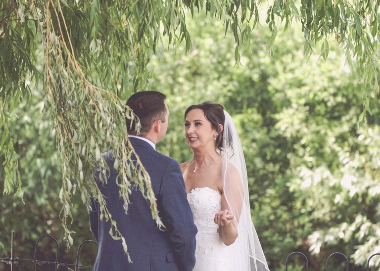 A bride looking at her groom taken through a willow tree on a wedding day at Little Green Wedding Barn photographed by suffolk wedding photographer Hayley Denston Photography