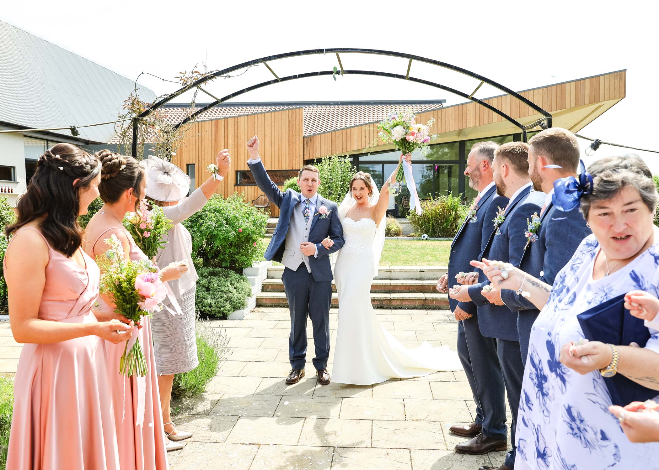 A bride and groom stood at the end of a confetti aisle holding their arms in the air on a wedding day at Little Green Wedding Barn photographed by suffolk wedding photographer Hayley Denston Photography