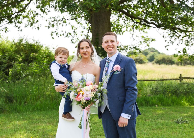 A bride holding her son on her hip with her groom on the otherside and an oak tree and fields behind them on a wedding day at Little Green Wedding Barn photographed by suffolk wedding photographer Hayley Denston Photography