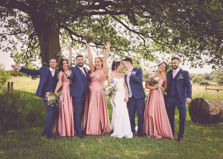 A bridal party in blues and pinks all pulling a pose for a group shot on a wedding day at Little Green Wedding Barn photographed by suffolk wedding photographer Hayley Denston Photography