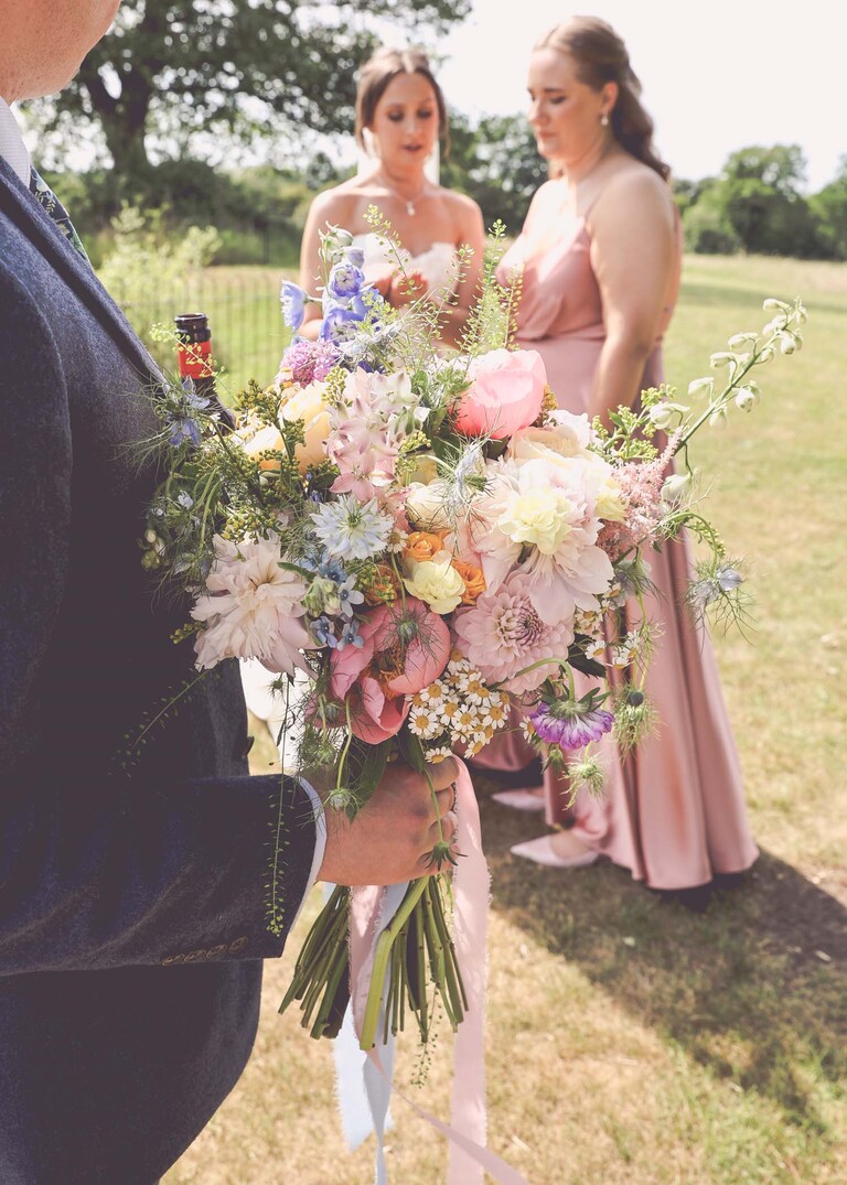 A close up of the grooms hand holding the brides bright floral bouquet on a wedding day at Little Green Wedding Barn photographed by suffolk wedding photographer Hayley Denston Photography