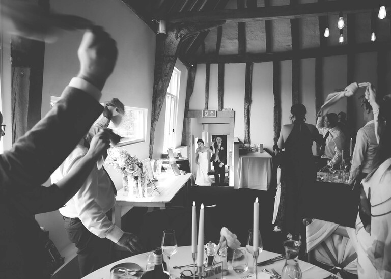A black and white photo of the bride and groom entering the wedding breakfast barn with guests waving napkins in the air on a wedding day at Little Green Wedding Barn photographed by suffolk wedding photographer Hayley Denston Photography
