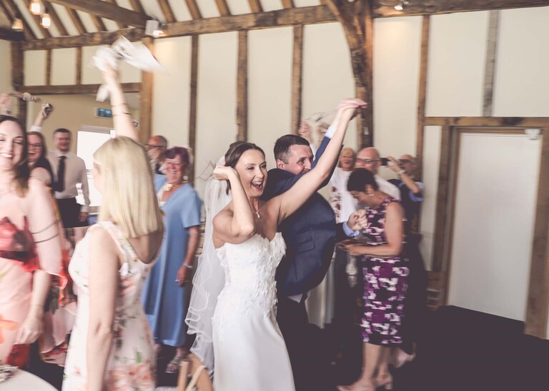 A bride and groom walking hand in hand with their arms in the air and guests holding up napkins on a wedding day at Little Green Wedding Barn photographed by suffolk wedding photographer Hayley Denston Photography
