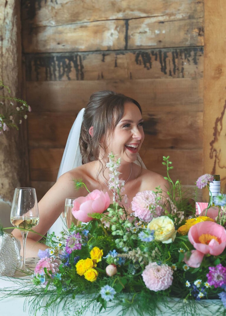 A bride with bright florals in front of her smiling during wedding speeches on a wedding day at Little Green Wedding Barn photographed by suffolk wedding photographer Hayley Denston Photography