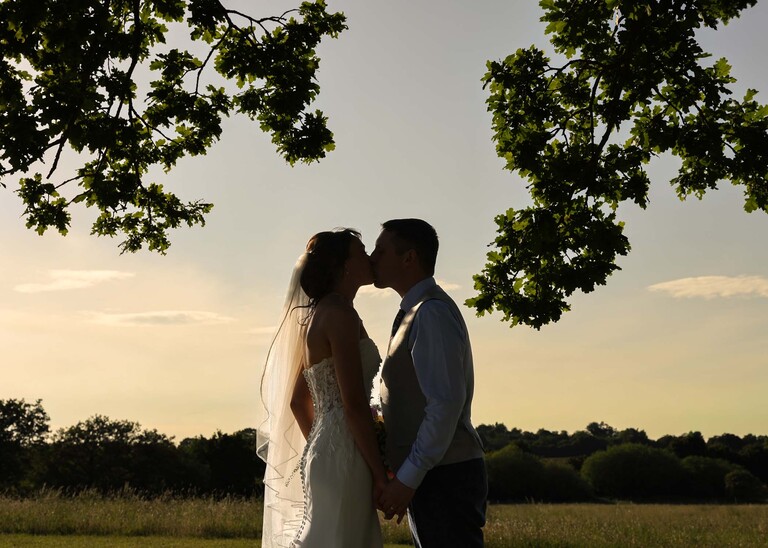 A silhouette of a bride and groom kissing at sunset under an oak tree on a wedding day at Little Green Wedding Barn photographed by suffolk wedding photographer Hayley Denston Photography