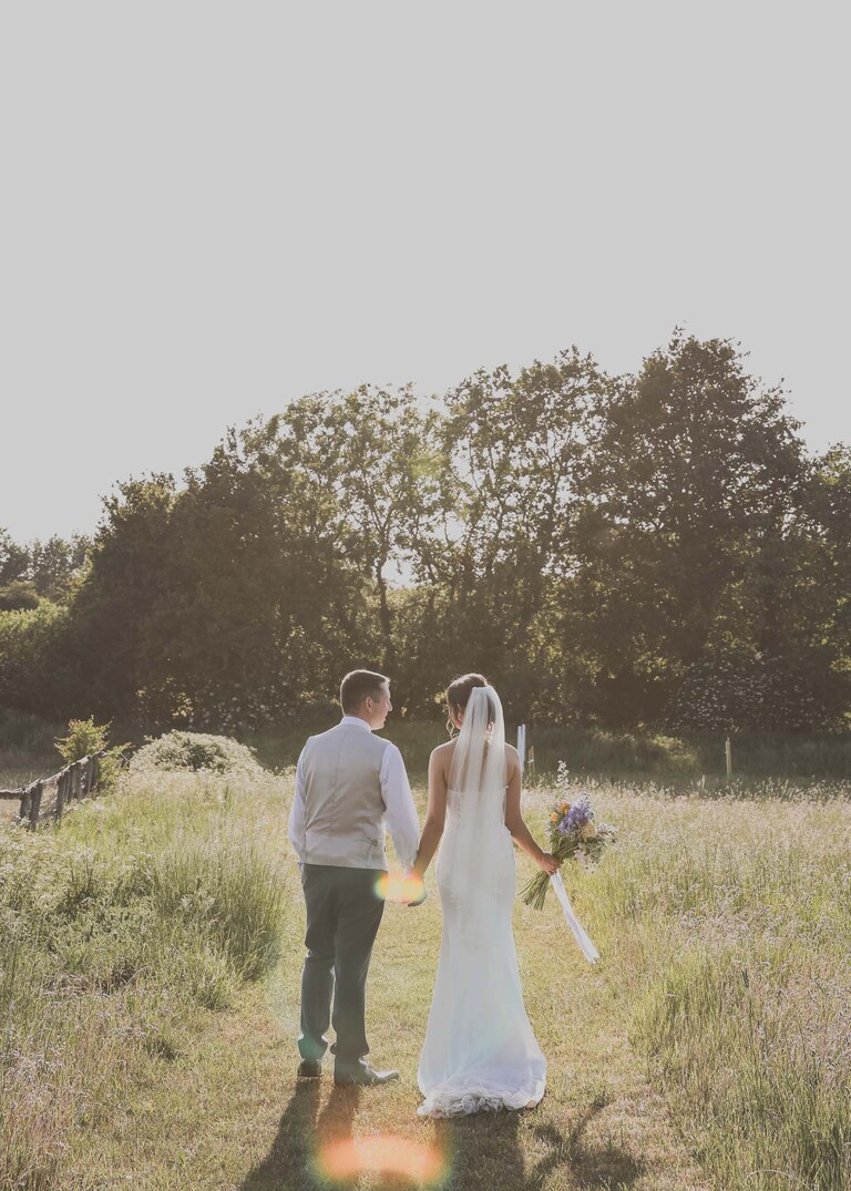 A bride and groom walking away through a meadow at golden hour on a wedding day at Little Green Wedding Barn photographed by suffolk wedding photographer Hayley Denston Photography