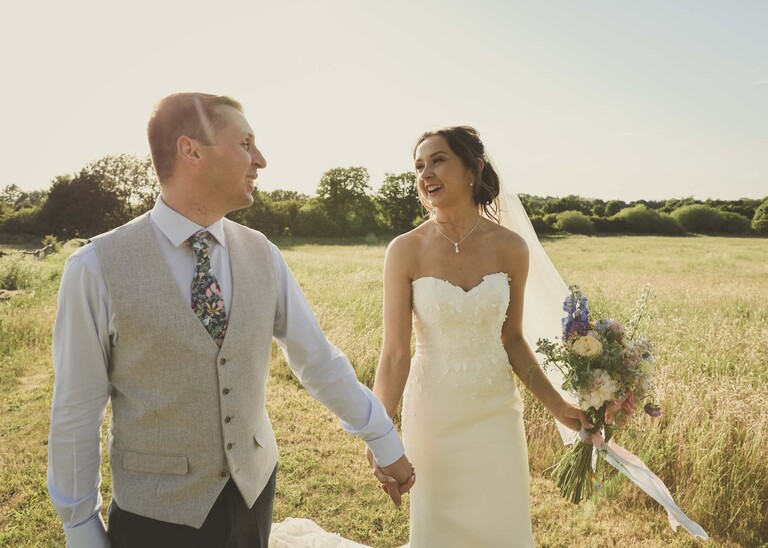 A bride and groom holding hands walking looking at each other during golden hour with fields and sun behind them on a wedding day at Little Green Wedding Barn photographed by suffolk wedding photographer Hayley Denston Photography