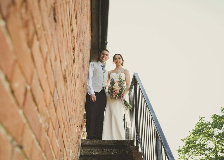 A bride and groom up the outdoor fire escape steps with red brick building on a wedding day at Little Green Wedding Barn photographed by suffolk wedding photographer Hayley Denston Photography