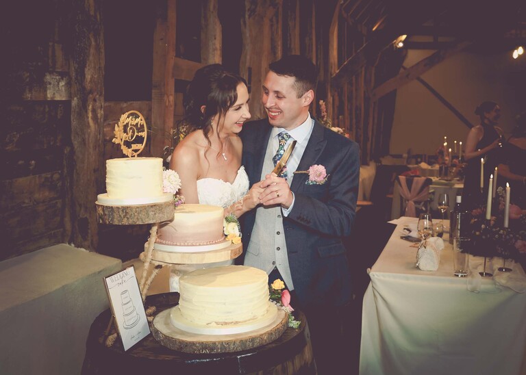 A bride and groom cutting their naked wedding cake laughing on a wedding day at Little Green Wedding Barn photographed by suffolk wedding photographer Hayley Denston Photography
