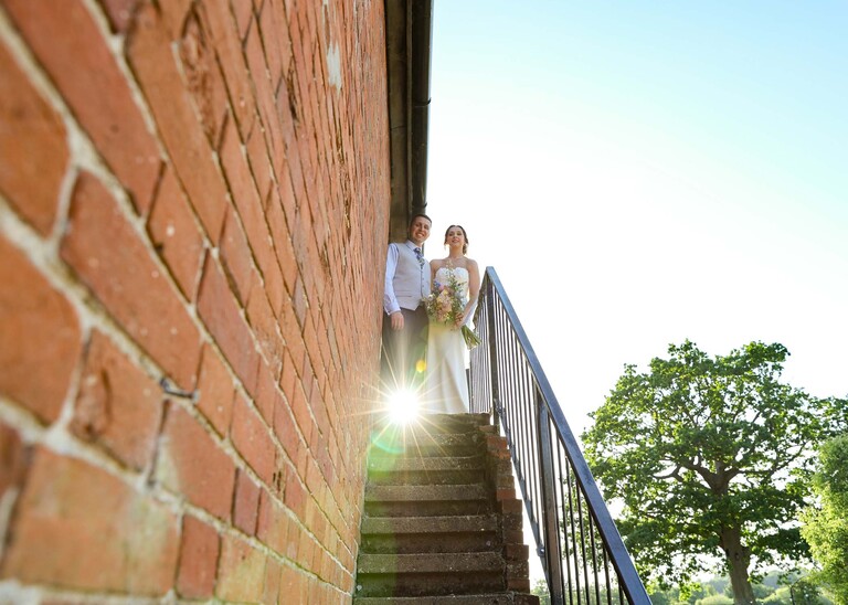 A bride and groom at the top of steps with the sun coming through and a red brick building on a wedding day at Little Green Wedding Barn photographed by suffolk wedding photographer Hayley Denston Photography
