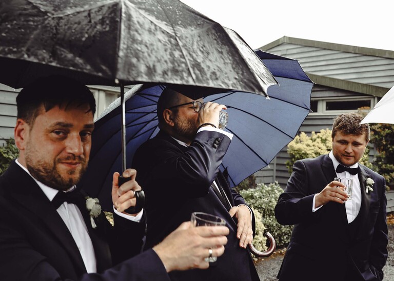 A groom and groomsmen all under umbrellas swigging from a hipflask before a wedding ceremony  on a wedding day at Hungarian Hall Estate photographed by suffolk wedding photographer Hayley Denston Photography