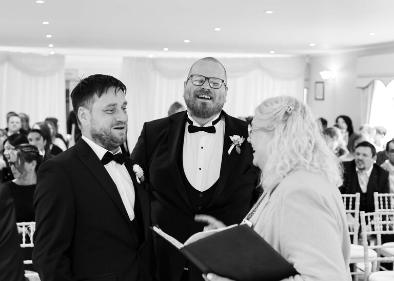 A black and white photo of a groom and bestman laughing with the registrar before the service  on a wedding day at Hungarian Hall Estate photographed by suffolk wedding photographer Hayley Denston Photography