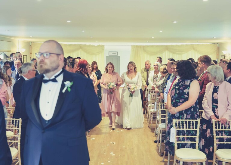 A bride and her sister walking down the aisle with the groom in the foreground  on a wedding day at Hungarian Hall Estate photographed by suffolk wedding photographer Hayley Denston Photography
