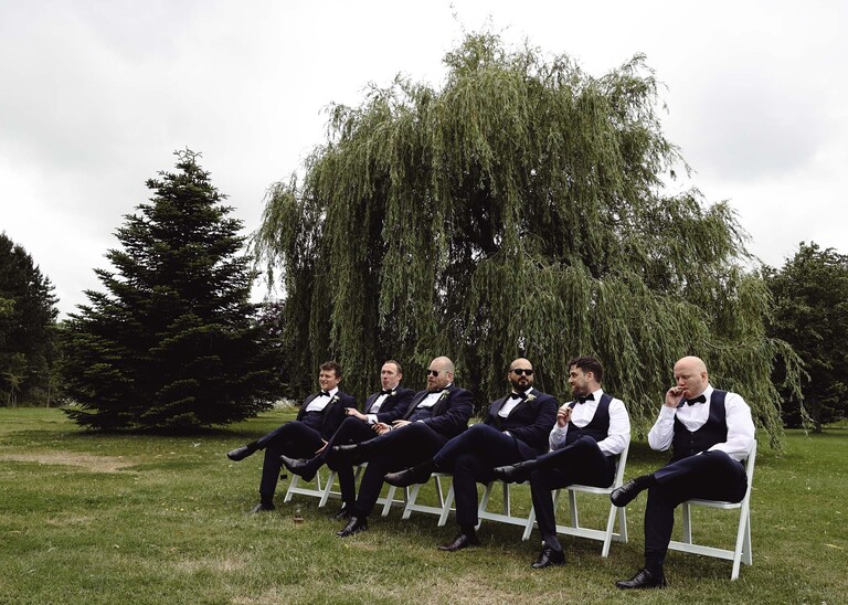 A groom and his groomsmen sat smoking cigars in front of a willow tree on a wedding day at Hungarian Hall Estate photographed by suffolk wedding photographer Hayley Denston Photography