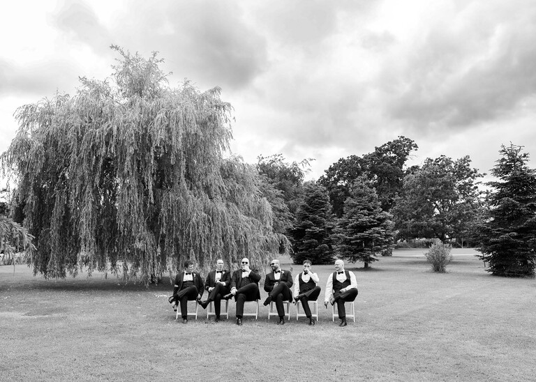 A black and white photo of a groom and groomsmen smoking cigars under a willow tree on a wedding day at Hungarian Hall Estate photographed by suffolk wedding photographer Hayley Denston Photography