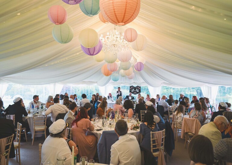 Wedding guests listening to speeches with colourful paper lanterns hanging from the roof of the marquee on a wedding day at Hungarian Hall Estate photographed by suffolk wedding photographer Hayley Denston Photography