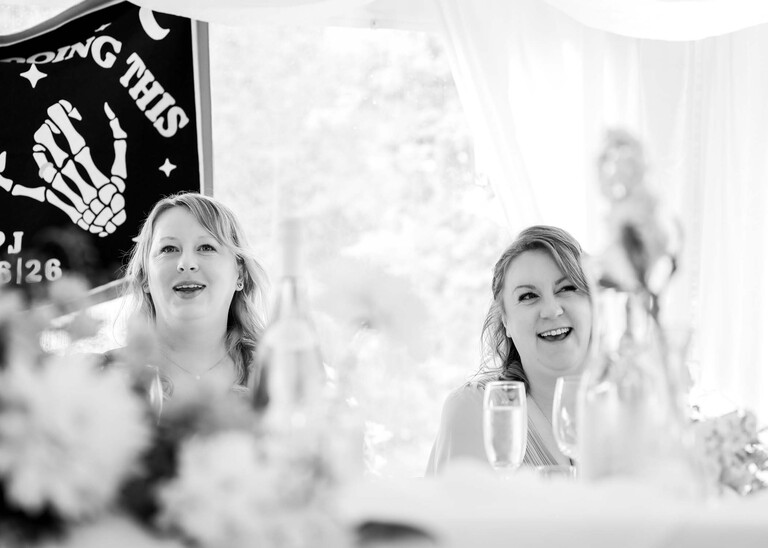 A black and white photo of a bride and her sister laughing during wedding speeches on a wedding day at Hungarian Hall Estate photographed by suffolk wedding photographer Hayley Denston Photography