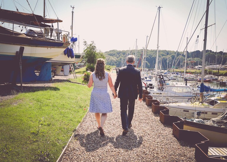 A bride and groom walking from behind through the yaughts in the marina on a wedding day in Woodbridge captured by Suffolk Wedding Photographer Hayley Denston Photography