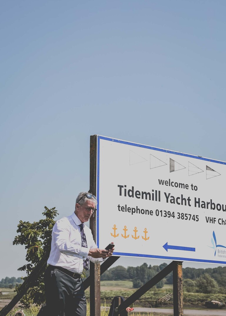 A groom leaning on the harbour sign at woodbridge marina on a wedding day in Woodbridge captured by Suffolk Wedding Photographer Hayley Denston Photography