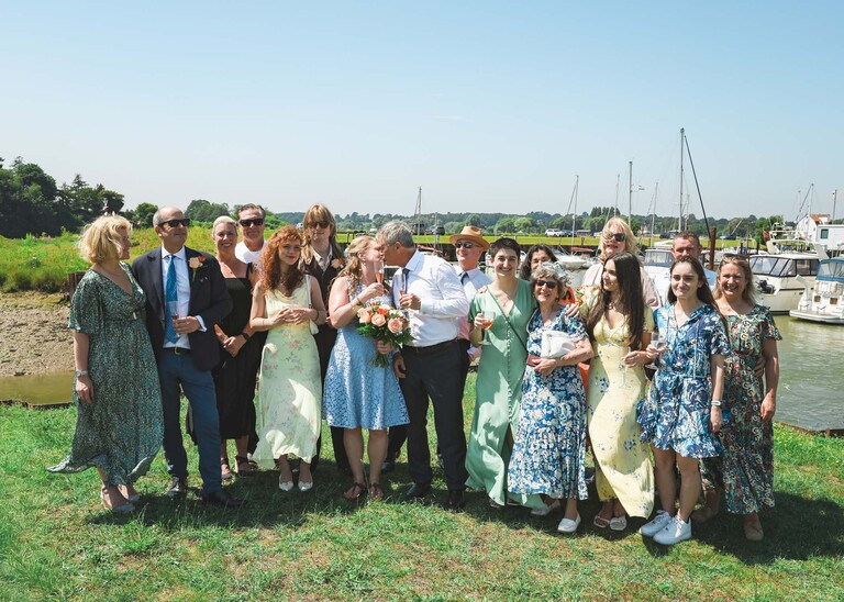 A group photo with the bride and groom kissing in the middle on the penisula at woodbridge marina on a wedding day in Woodbridge captured by Suffolk Wedding Photographer Hayley Denston Photography