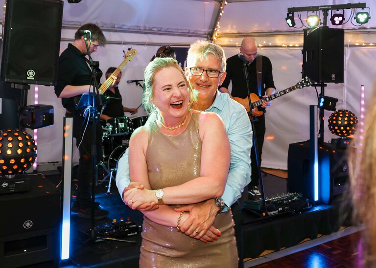 A groom with his arms round the bride both laughing and dancing with the band in the background at an evening wedding reception at the Unruly Pig in Suffolk captured by Suffolk Wedding Photographer Hayley Denston Photography
