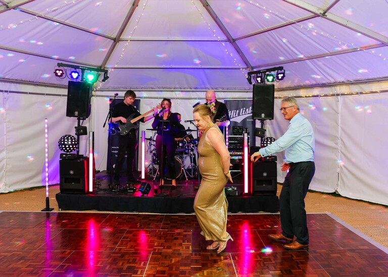 A bride and groom having their first dance with the band behind them in a lit marquee in gardens for an evening wedding reception at the Unruly Pig in Suffolk captured by Suffolk Wedding Photographer Hayley Denston Photography