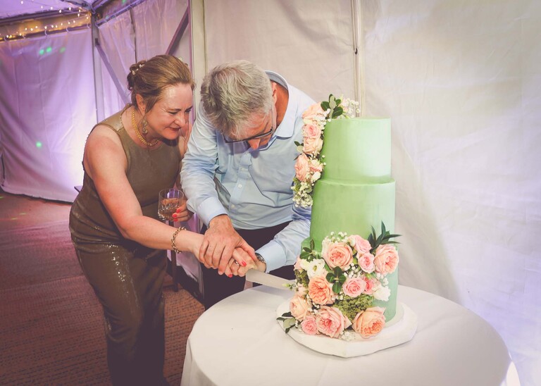 A bride and groom cutting their green wedding cake in a lit marquee in gardens at an evening wedding reception at the Unruly Pig in Suffolk captured by Suffolk Wedding Photographer Hayley Denston Photography